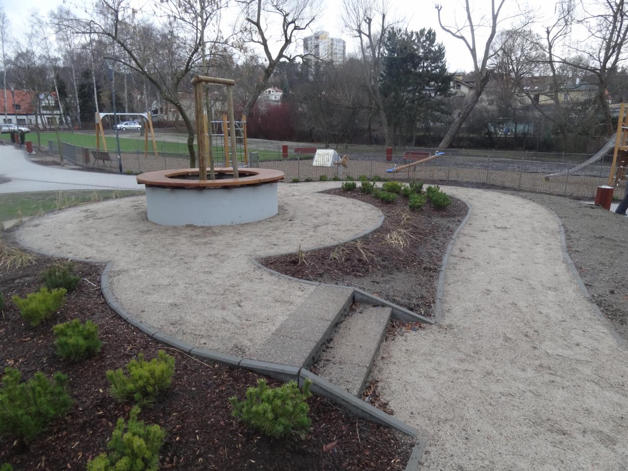 Wykończona ścieżka z kostki brukowej na placu zabaw, otoczona zielenią i elementami drewnianymi. Widoczne schody z kostki i obrzeża. Całość tworzy estetyczną przestrzeń.