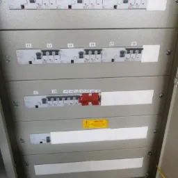 Szczegółowe ujęcie rozdzielnicy elektrycznej z numerowanymi wyłącznikami, niektóre zabezpieczone czerwoną taśmą, oraz żółtą naklejką ostrzegawczą.