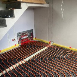 PRUNAM Ryszard Maciąg - Montaż ogrzewania podłogowego: widok na rozłożone rury grzewcze w systemie podłogowym, z widoczną rozdzielnią i elementami instalacji pod schodami w surowym wnętrzu.