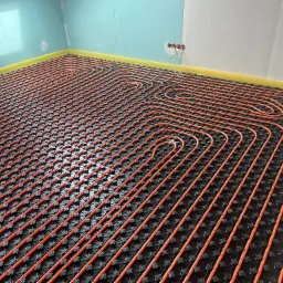 Pomarańczowe rury ogrzewania podłogowego ułożone na czarnej macie z wypustkami w nowym budynku z surowymi ścianami, widoczny fragment poziomicy.