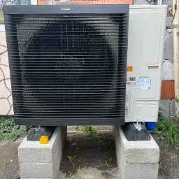 Zewnętrzna jednostka pompy ciepła Daikin zainstalowana na betonowych bloczkach, widoczna tabliczka znamionowa i elementy instalacji przy ścianie budynku i drewnianym ogrodzeniu.