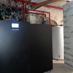 Nowoczesny, czarny piec z wyświetlaczem Kostrzewa w piwnicy z odsłoniętymi instalacjami i surowym, betonowym filarem.