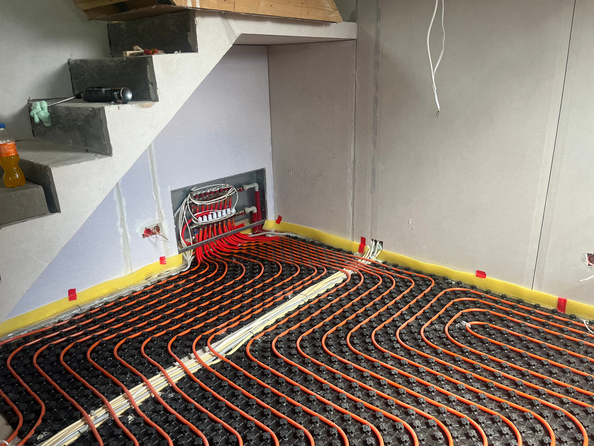 Montaż ogrzewania podłogowego: widok na rozłożone rury grzewcze w systemie podłogowym, z widoczną rozdzielnią i elementami instalacji pod schodami w surowym wnętrzu.