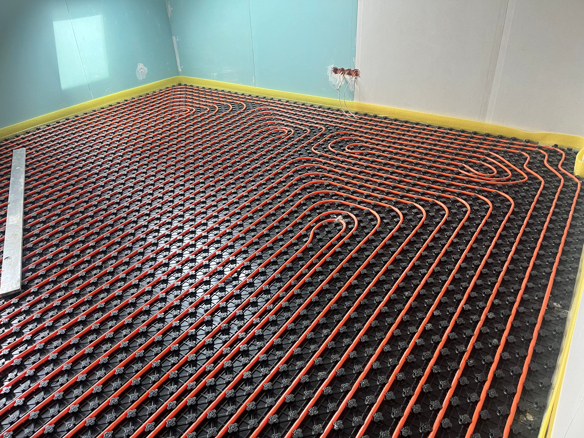 Pomarańczowe rury ogrzewania podłogowego ułożone na czarnej macie z wypustkami w nowym budynku z surowymi ścianami, widoczny fragment poziomicy.