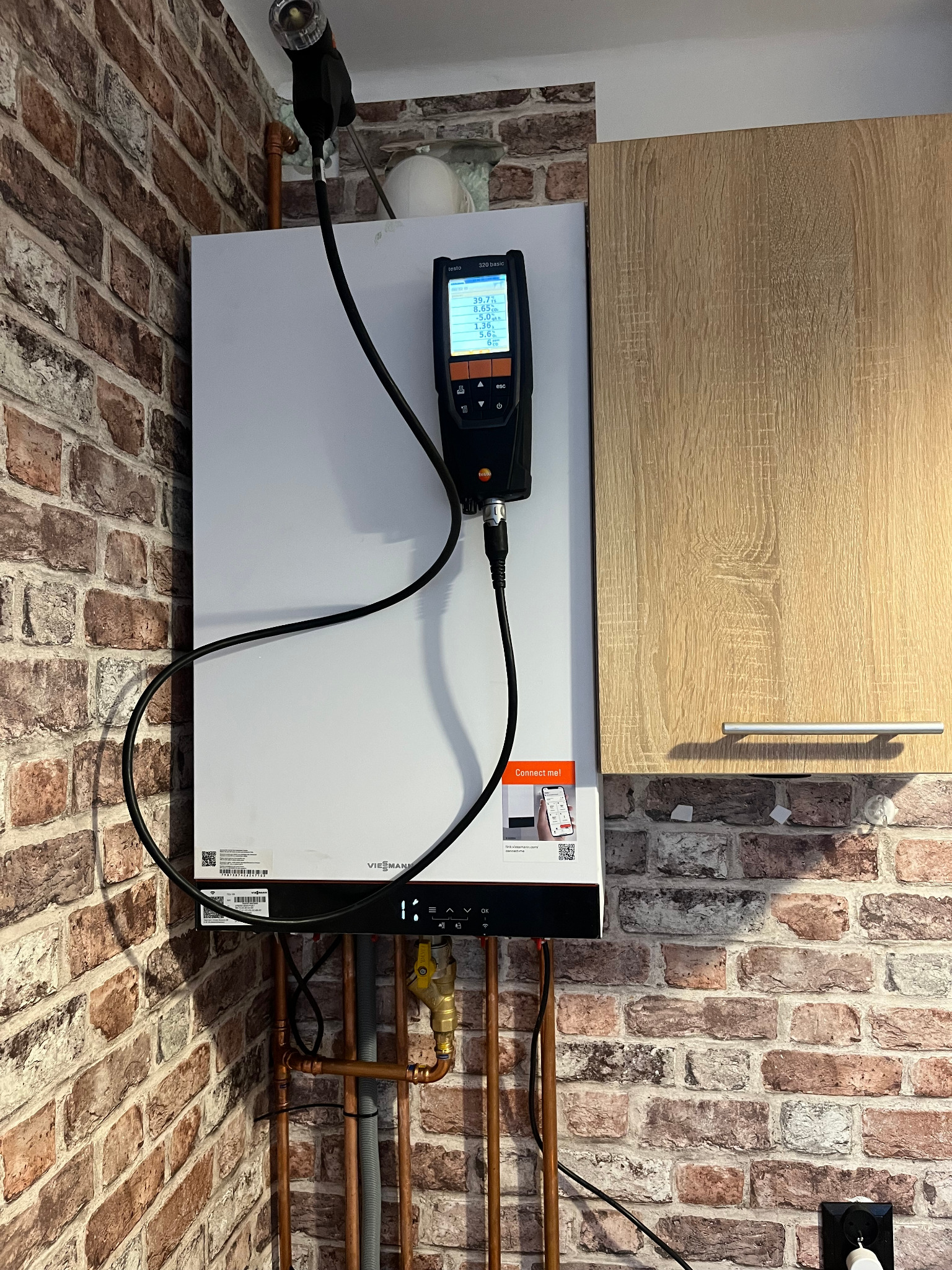 Biały piec gazowy Viessmann zamontowany na ścianie z cegieł, obok szafki kuchennej, podłączony do analizatora spalin Testo 320 basic podczas pomiarów.
