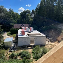 Wznoszenie ścian parterowego domu z białych bloczków, widoczne materiały izolacyjne na stropie, w tle zalesiony teren i zabudowania.