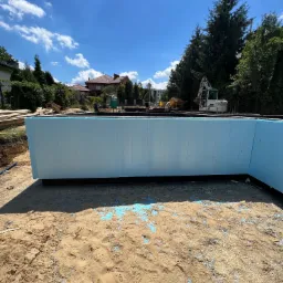 Niebieska izolacja termiczna fundament&oacute;w budynku w trakcie montażu, z widocznym wykopem, piaskiem i sprzętem budowlanym w tle.