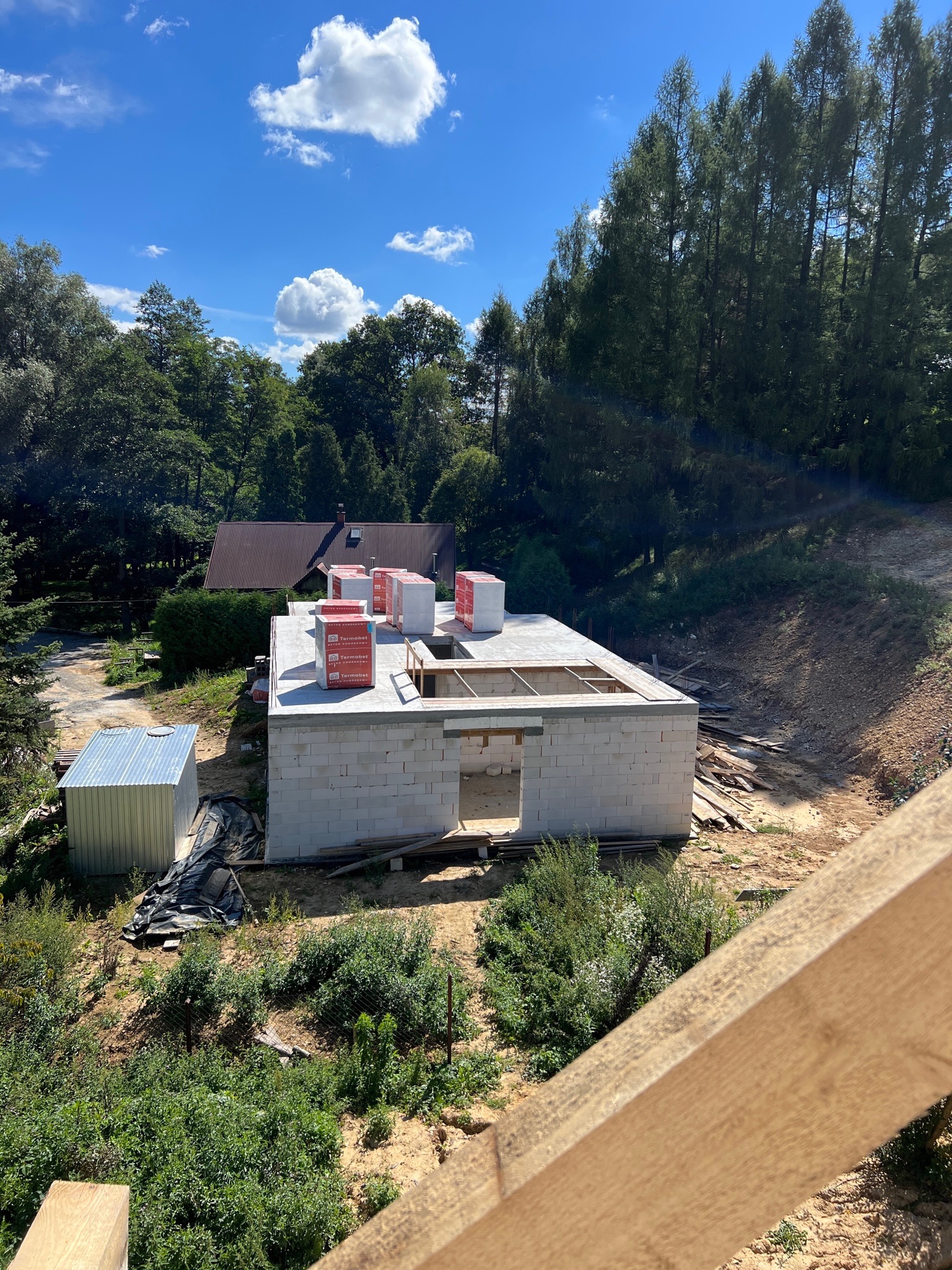 Wznoszenie ścian parterowego domu z białych bloczków, widoczne materiały izolacyjne na stropie, w tle zalesiony teren i zabudowania.