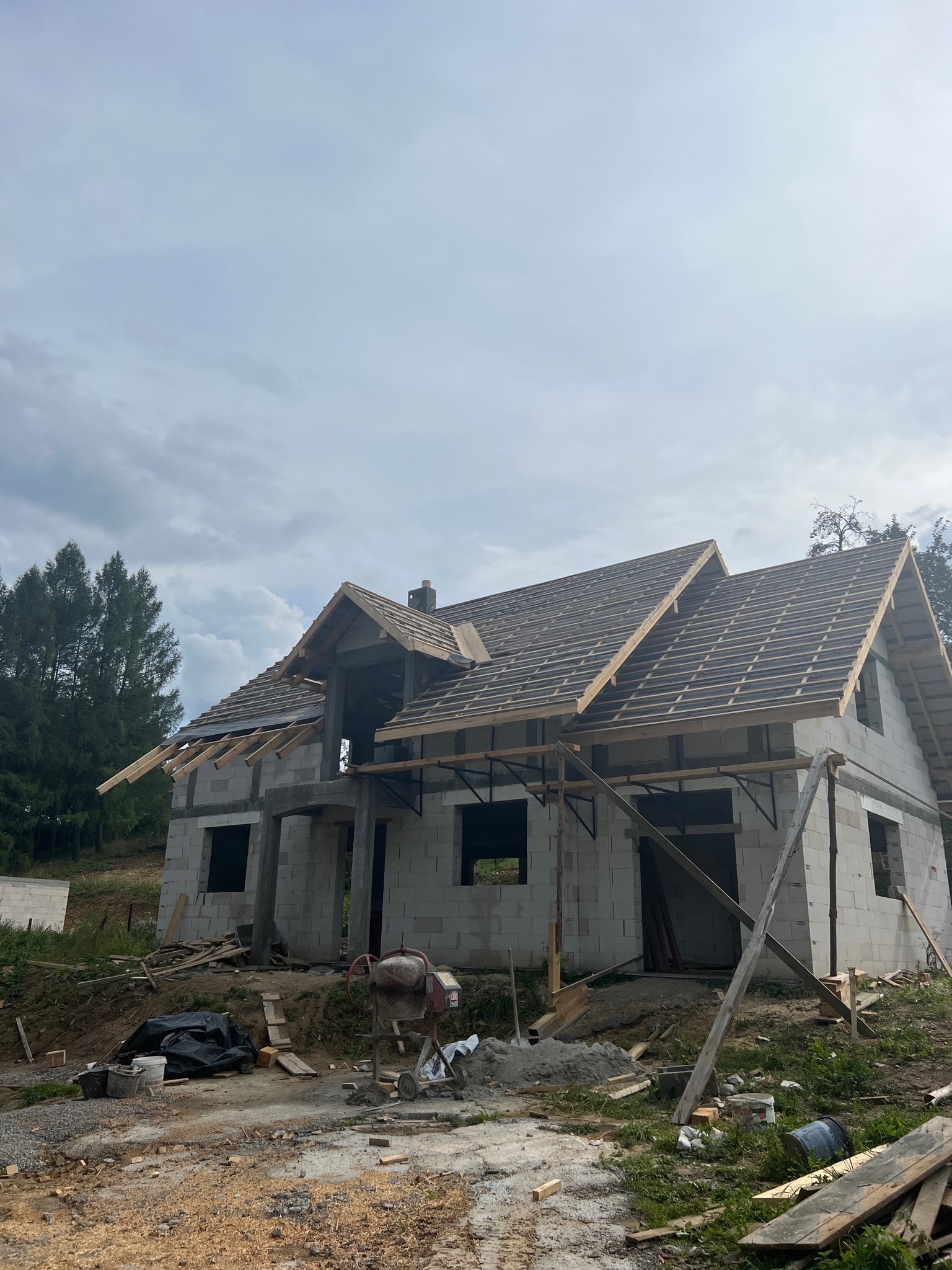 Surowy stan domu z betonu komórkowego w trakcie budowy, widoczna więźba dachowa, betonowa podmurówka i mieszalnik do zaprawy na placu budowy.