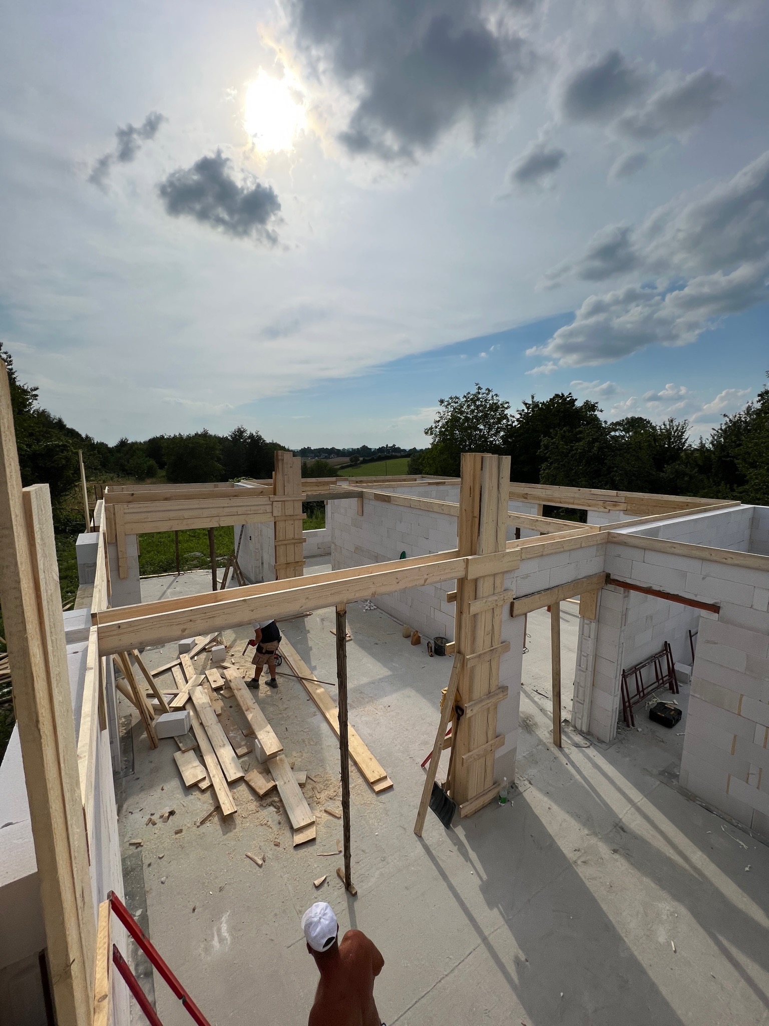 Budowa domu w trakcie realizacji: wzniesione ściany z bloczków, drewniane elementy konstrukcyjne, widok z góry na plac budowy w słoneczny dzień, pracownik w oddali.