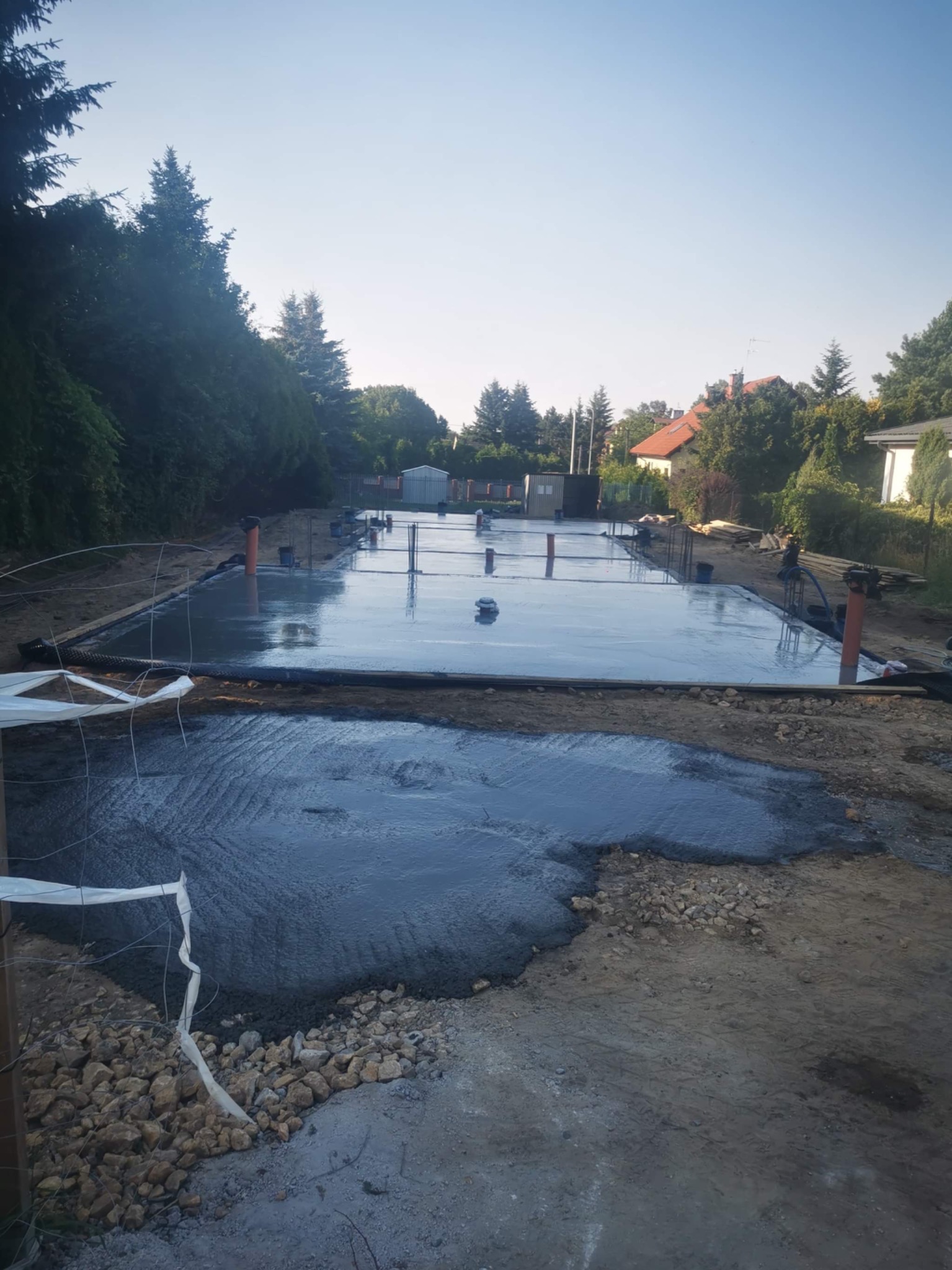 Świeżo wylany beton na fundamentach domu jednorodzinnego, widoczne wystające rury i zbrojenie, częściowo zalane kamienie w wykopie.