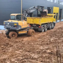 Żółta koparka Volvo EW140C nabiera ziemię do żółtej ciężarówki MAN na błotnistym terenie budowy z szaro-żółtym budynkiem w tle.