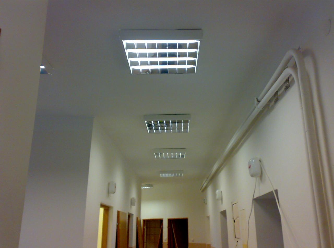 Długi korytarz oświetlony szeregiem kwadratowych lamp sufitowych z rastrem. Widoczne rury instalacyjne wzdłuż ściany, drzwi po obu stronach korytarza.