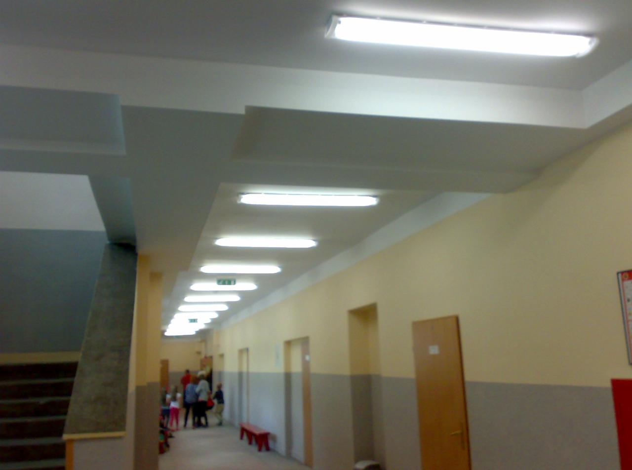 Długi korytarz oświetlony szeregiem lamp fluorescencyjnych, z widocznymi schodami po lewej i osobami w oddali. Ściany dwukolorowe, kremowo-szare.