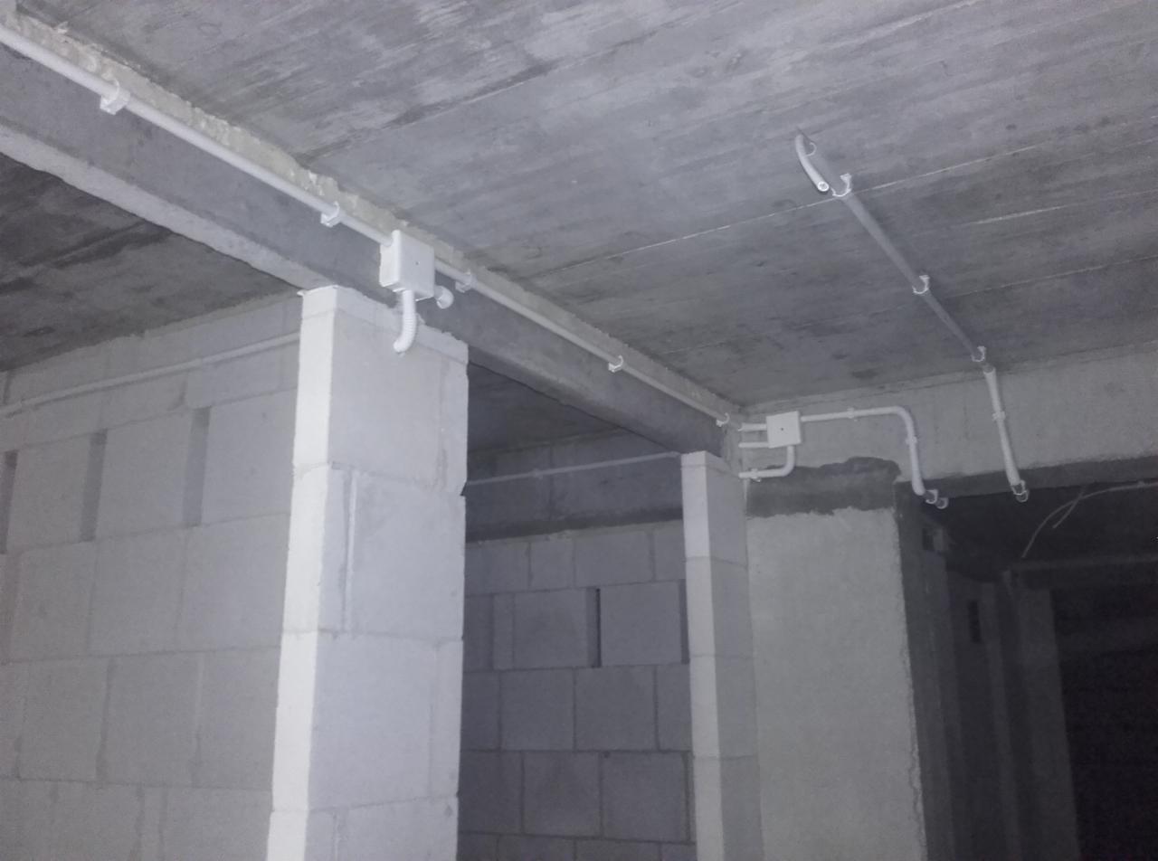 Surowy strop z widoczną, białą instalacją elektryczną natynkową, prowadzoną wzdłuż betonowych belek. Białe kable i puszki na tle szarego betonu.