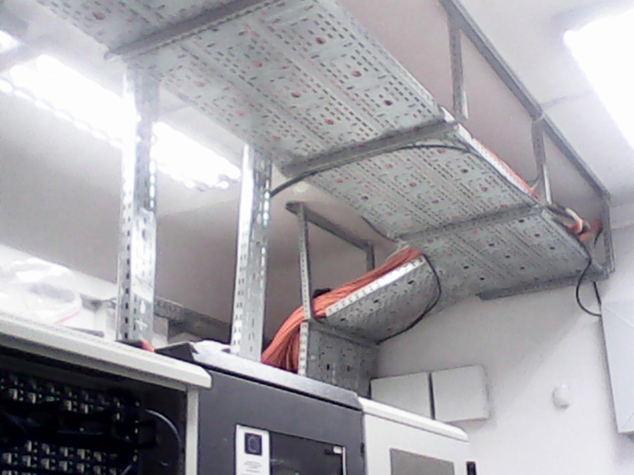 Metalowe korytka kablowe pod sufitem, prowadzące kable w budynku. Widoczne elementy szafy serwerowej.
