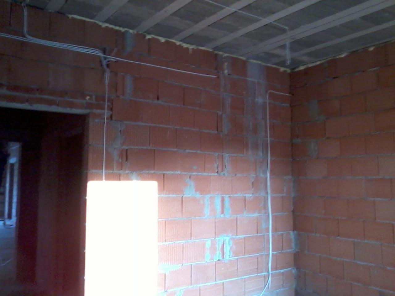 Surowa ściana z cegły z widoczną instalacją elektryczną w białych rurkach, poprowadzoną wzdłuż sufitu i pionowo po ścianie, w trakcie prac wykończeniowych.