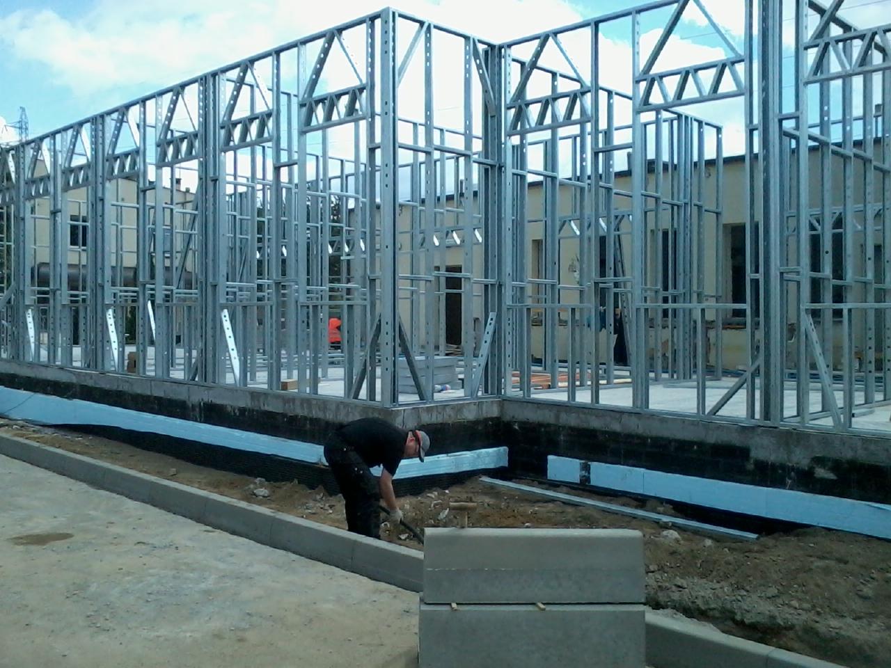 Budowa domu: szkielet stalowy na fundamencie, ocieplenie, robotnik z łopatą. Widoczna konstrukcja budynku w trakcie wznoszenia, z elementami izolacji termicznej.