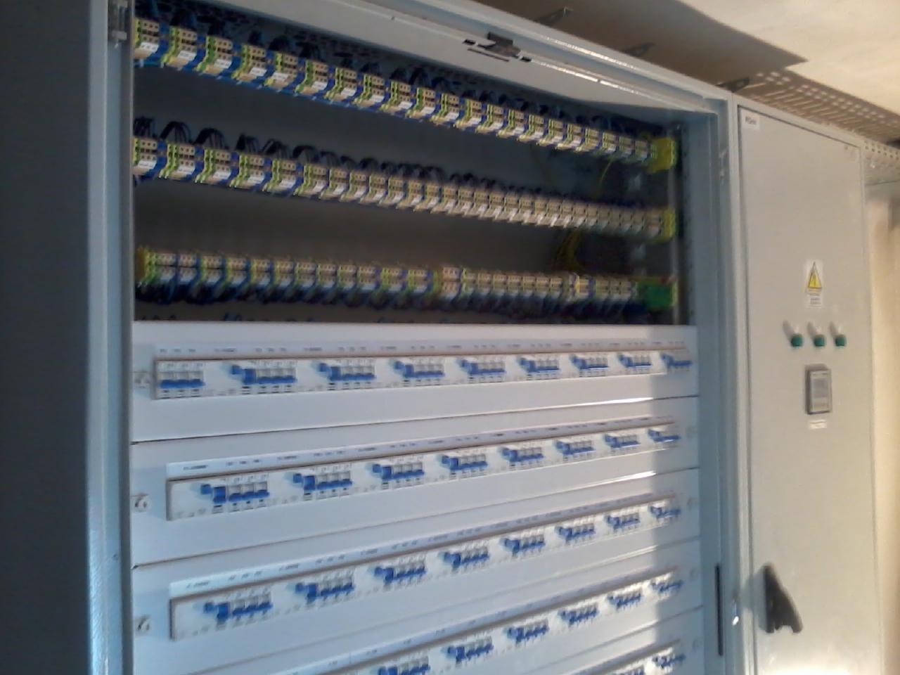 Otwarta, szara szafa elektryczna z widocznymi bezpiecznikami i przewodami. Górne rzędy z przewodami w kolorowych izolacjach, dolne z bezpiecznikami. Widoczny znak ostrzegawczy.