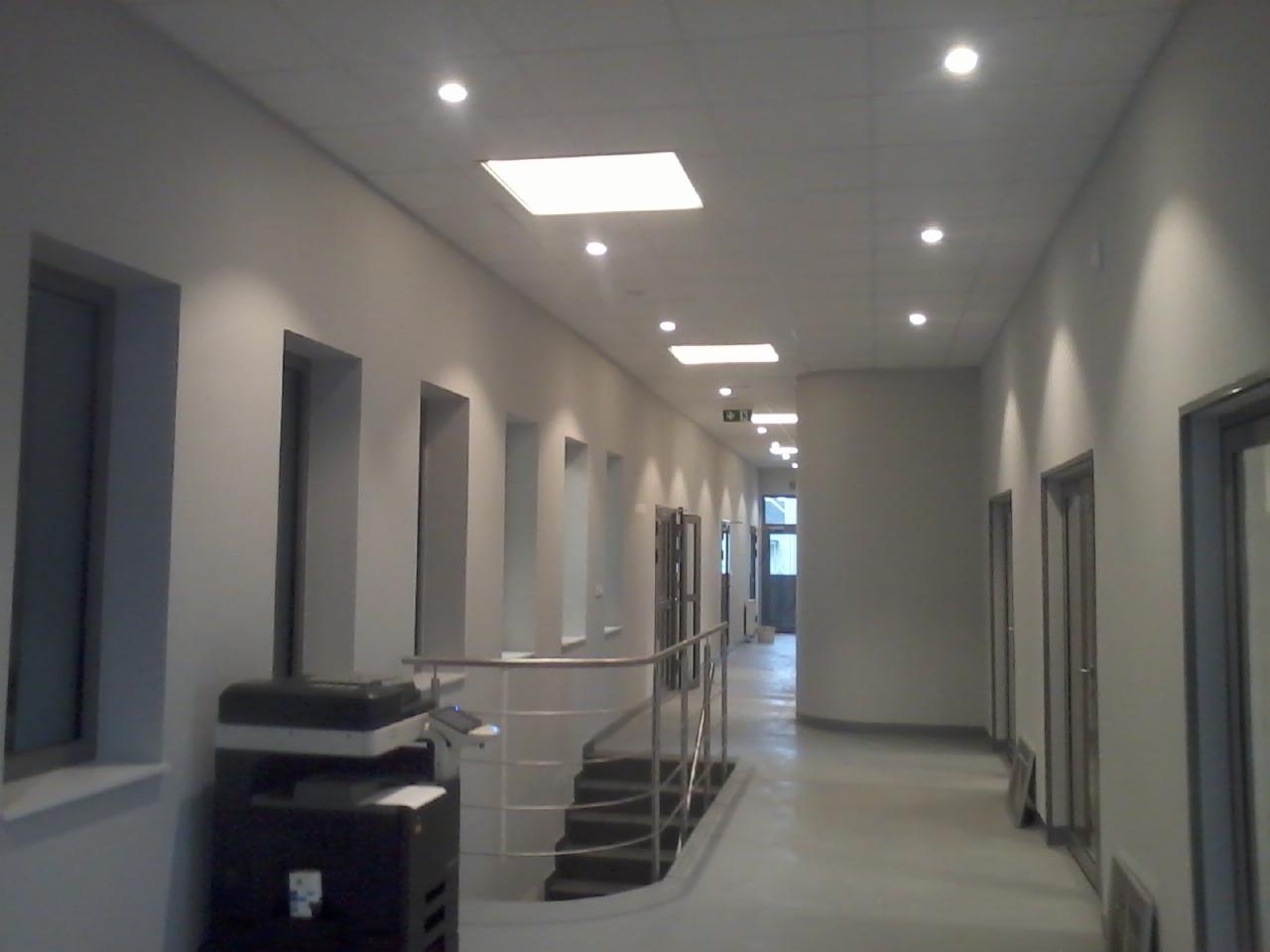 Jasny korytarz biurowy z równomiernie rozmieszczonymi, kwadratowymi i okrągłymi lampami sufitowymi. Na ścianie widoczny czarny automat biurowy.