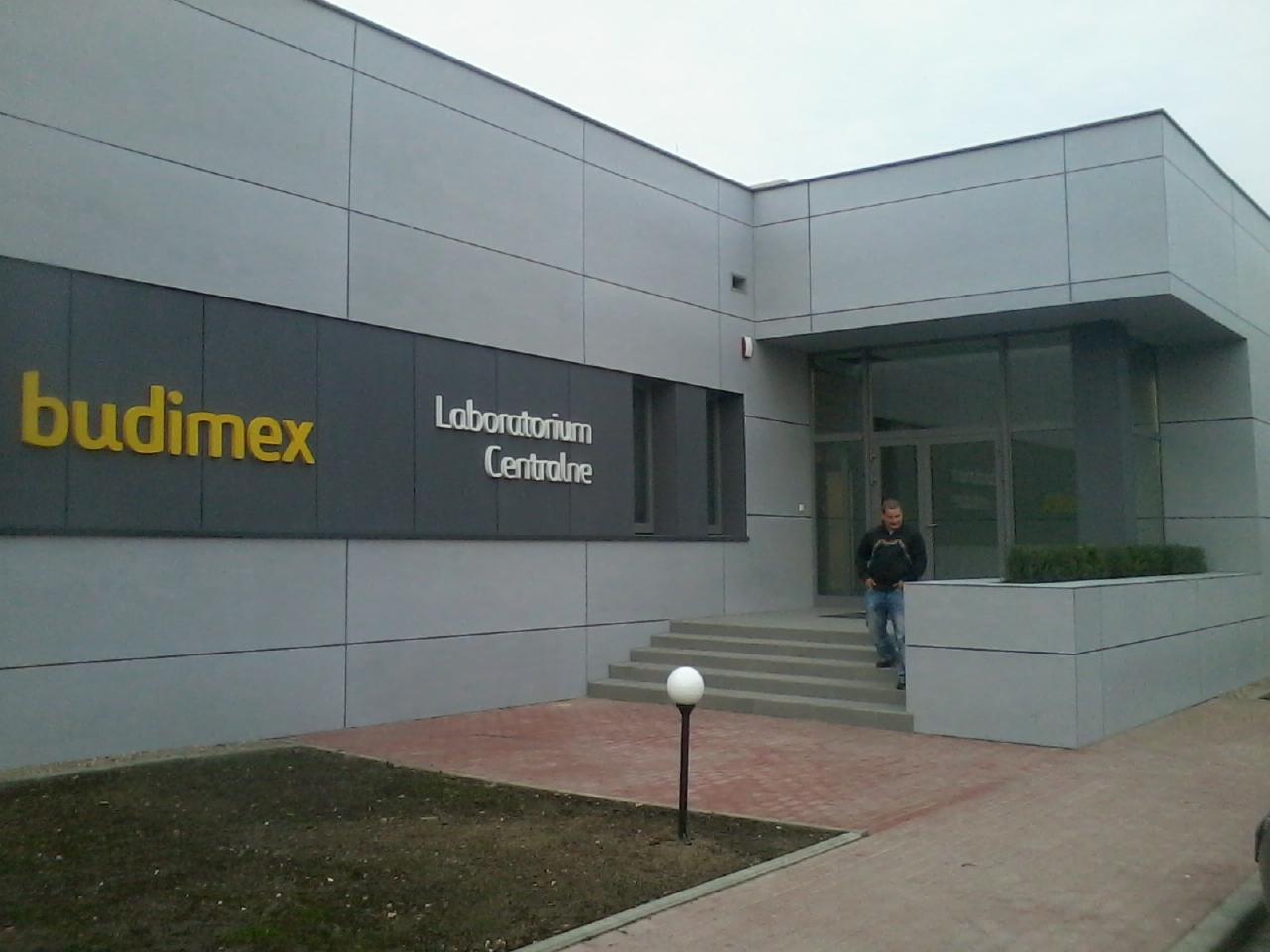 Budynek z napisem 'budimex Laboratorium Centralne', schody wejściowe i mężczyzna wchodzący do budynku. Szara elewacja z panelami.