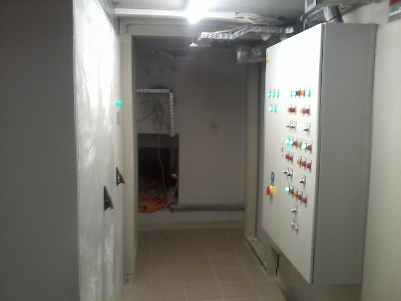 Szara szafa sterownicza z włącznikami i diodami LED w korytarzu, widoczne przewody i instalacje pod sufitem. Oświetlenie sufitowe.