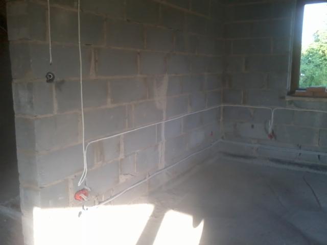 Surowy pokój z szarego betonu komórkowego, z widocznymi białymi przewodami elektrycznymi w ścianach i podłodze, okno z widokiem na zieleń.