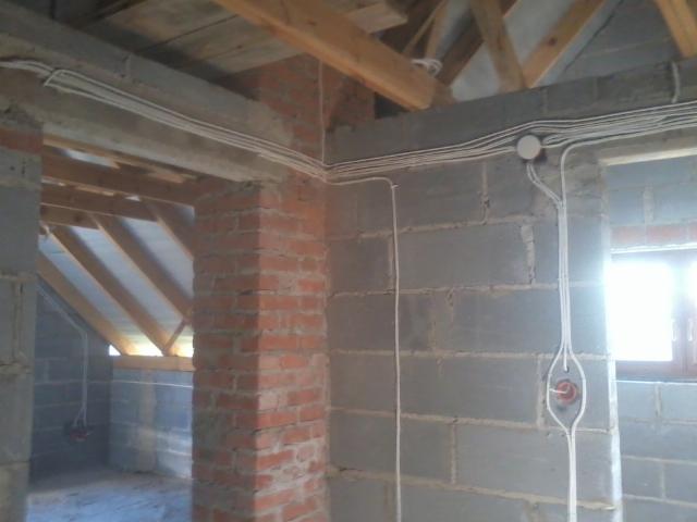 Instalacja elektryczna w surowym budynku: przewody prowadzone po ścianach z betonu komórkowego i cegły, widoczne elementy poddasza z drewnianą konstrukcją.