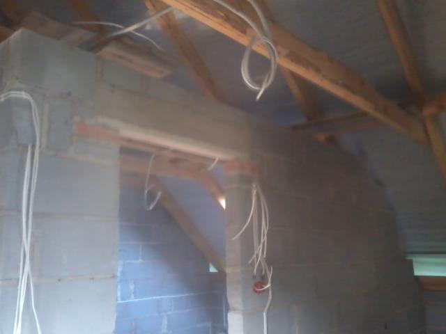 Surowa ściana z bloczków z widocznymi przewodami elektrycznymi, drewniana konstrukcja dachu w tle, wnętrze w trakcie prac wykończeniowych.