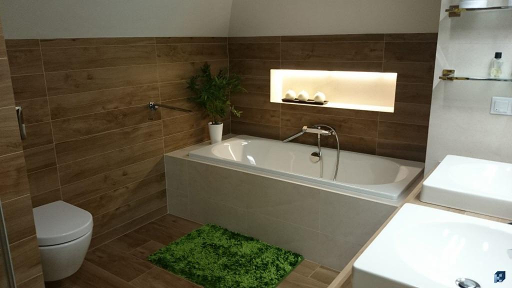 Nowoczesna łazienka z wanną obudowaną płytkami, drewnopodobne płytki na ścianach i podłodze, dwie białe umywalki i zielony dywanik.