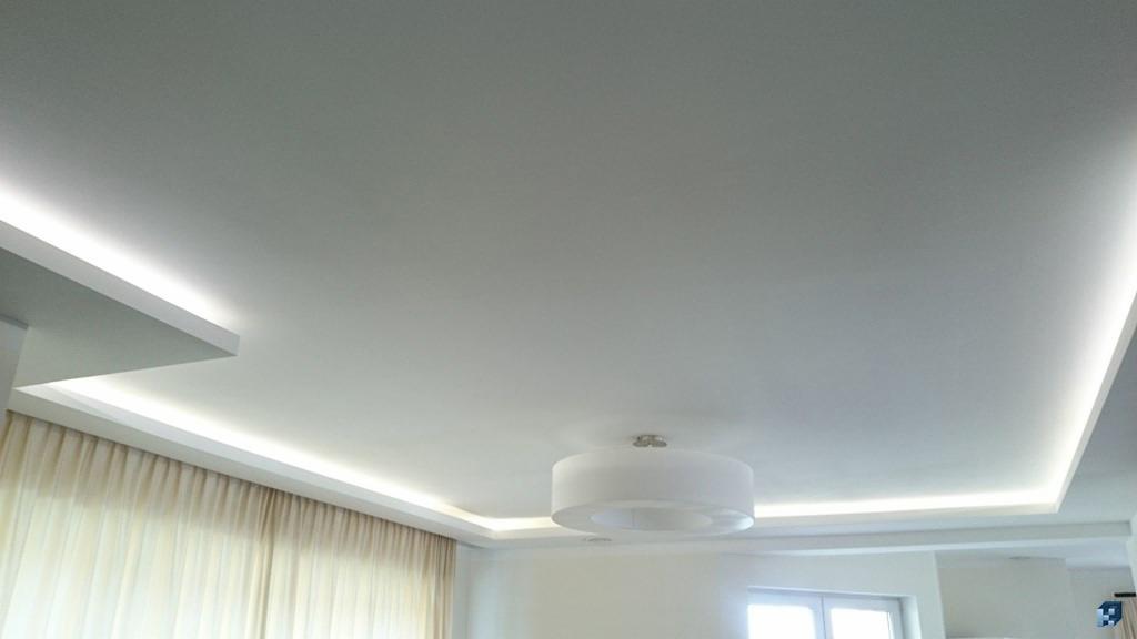 Nowoczesny sufit podwieszany z oświetleniem LED wzdłuż krawędzi. Okrągła lampa sufitowa w centrum, zasłony w tle. Minimalistyczny design i subtelne oświetlenie.