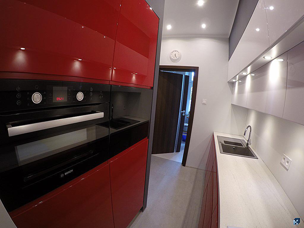 Nowoczesna kuchnia z czerwonymi szafkami, czarnym piekarnikiem Bosch i białymi szafkami górnymi z oświetleniem punktowym. Widok na otwarte drzwi do innego pomieszczenia.