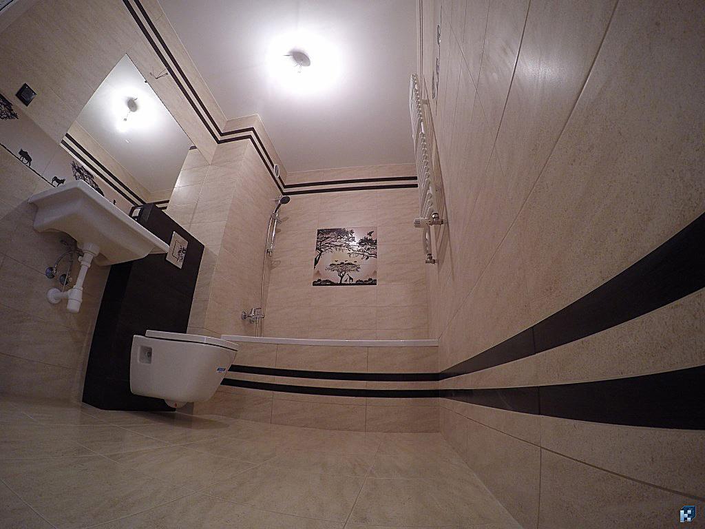 Wykończona łazienka w beżowych płytkach z czarnymi pasami dekoracyjnymi. Widoczna umywalka, toaleta, wanna i grzejnik drabinkowy. Na ścianie dekoracyjny panel z motywem afrykańskim.