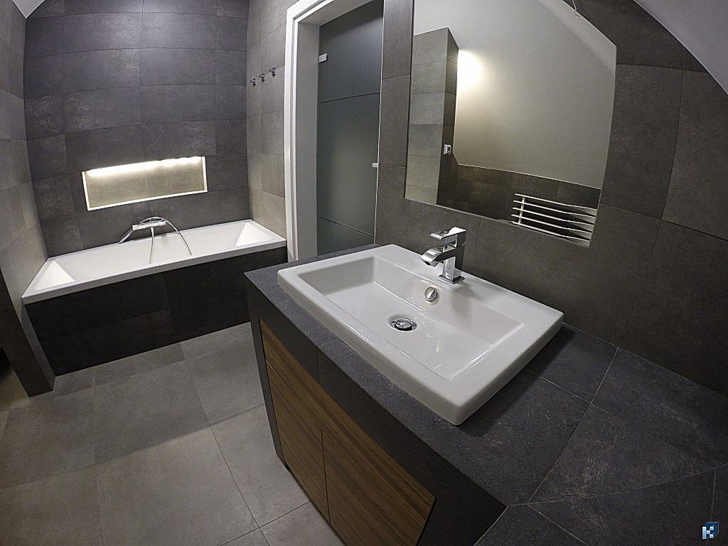 Nowoczesna łazienka w szarościach: wanna z podświetleniem, umywalka na szafce z drewnianymi frontami, duże lustro i ciemne płytki. Minimalistyczny design i funkcjonalność.