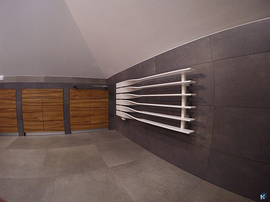 Nowoczesna łazienka z szarą, dużą płytką, drewnianymi szafkami i dekoracyjnym, białym grzejnikiem. Ukośny sufit dodaje unikalnego charakteru wnętrzu.