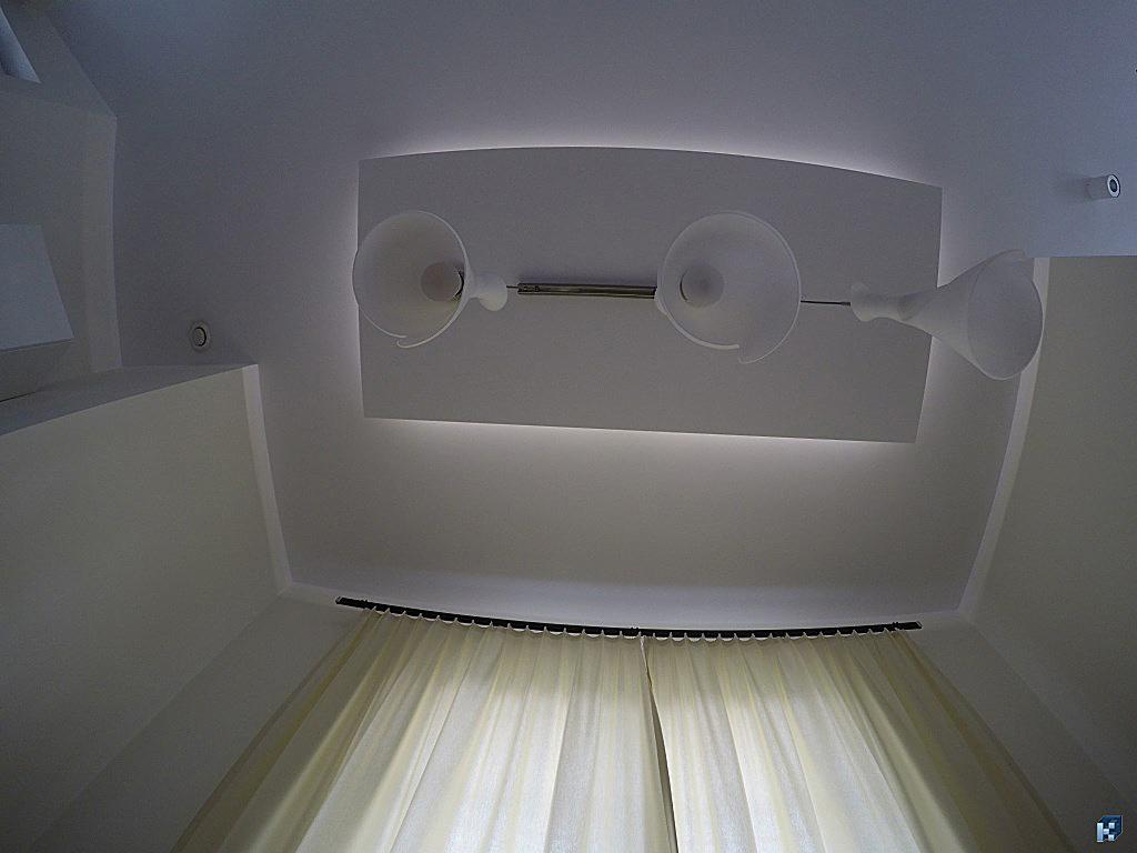 Nowoczesny sufit podwieszany z oświetleniem punktowym i delikatnymi zasłonami w tle. Białe, minimalistyczne wnętrze z akcentem na detale wykończeniowe.