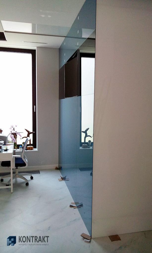 Szklana ściana działowa z widocznymi elementami mocującymi na podłodze, zabezpieczona kartonem, w jasnym biurze z dużym oknem i biurkiem z krzesłem.