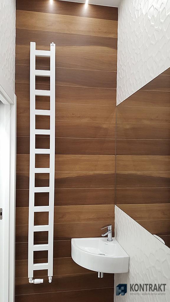 Nowoczesna łazienka z drewnopodobnymi płytkami, białą umywalką narożną i grzejnikiem drabinkowym, oświetlona punktowo, z fragmentem dekoracyjnej, białej ściany o trójwymiarowej fakturze.