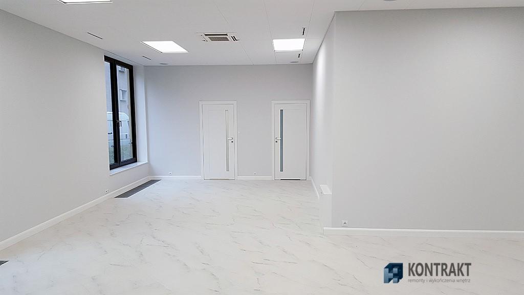Jasne, świeżo pomalowane biuro z marmurową podłogą, białymi drzwiami z przeszkleniami i czarną ramą okienną, widoczne kratki wentylacyjne sufitowe.
