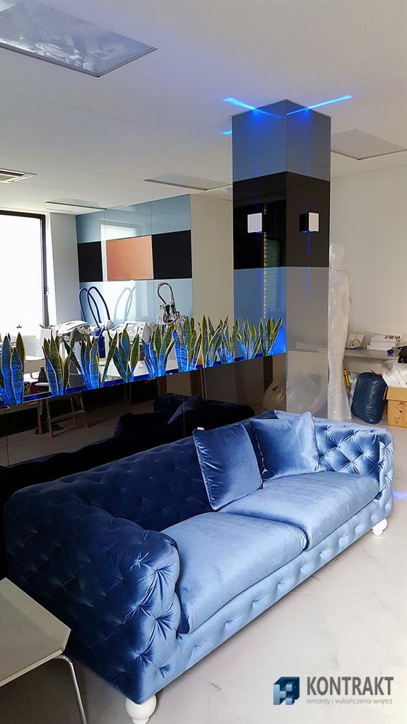 Niebieska sofa chesterfield z weluru na marmurowej podłodze, na tle lustrzanej ściany z podświetlanymi roślinami i kolumną z niebieskim oświetleniem w nowoczesnym biurze.