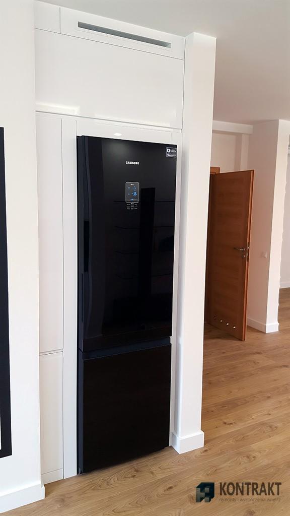 Nowoczesna kuchnia z czarną lodówką Samsung w zabudowie, białe szafki, drewniana podłoga i częściowo otwarta drewniana drzwi w tle.