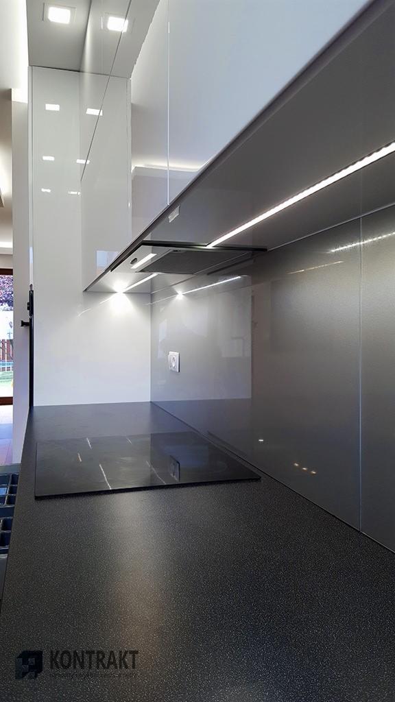 Nowoczesna kuchnia z białymi szafkami na wysoki połysk, szarym panelem ściennym z oświetleniem LED podszafkowym i czarnym blatem z wbudowaną płytą indukcyjną.