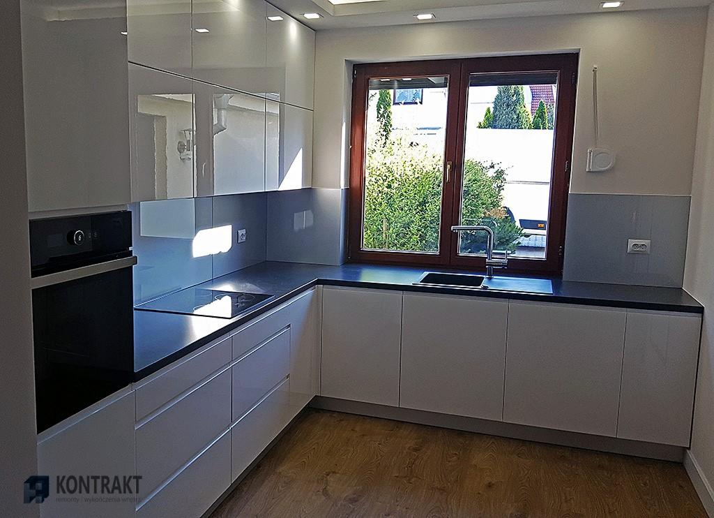 Nowoczesna kuchnia z białymi, lakierowanymi szafkami, ciemnym blatem i drewnianą podłogą, widok na zieleń za oknem.