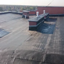 Płaski dach pokryty czarną membraną bitumiczną z widocznymi kominami wentylacyjnymi i obróbkami blacharskimi, w tle zabudowa miejska i drzewa.