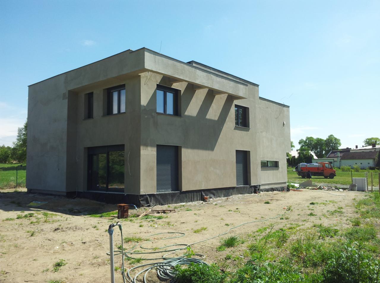 Surowa elewacja dwukondygnacyjnego domu szkieletowego w trakcie budowy, widoczne okna z szarymi roletami, teren wokół domu jeszcze niezagospodarowany, w tle zaparkowany pomarańczowy samochód...