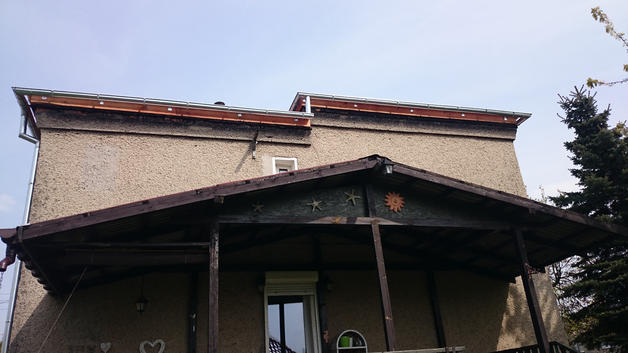 Dwupiętrowy dom z beżową elewacją i nowymi metalowymi rynnami, widocznymi na krawędzi dachu, z werandą ozdobioną drewnianymi gwiazdami i słońcem.