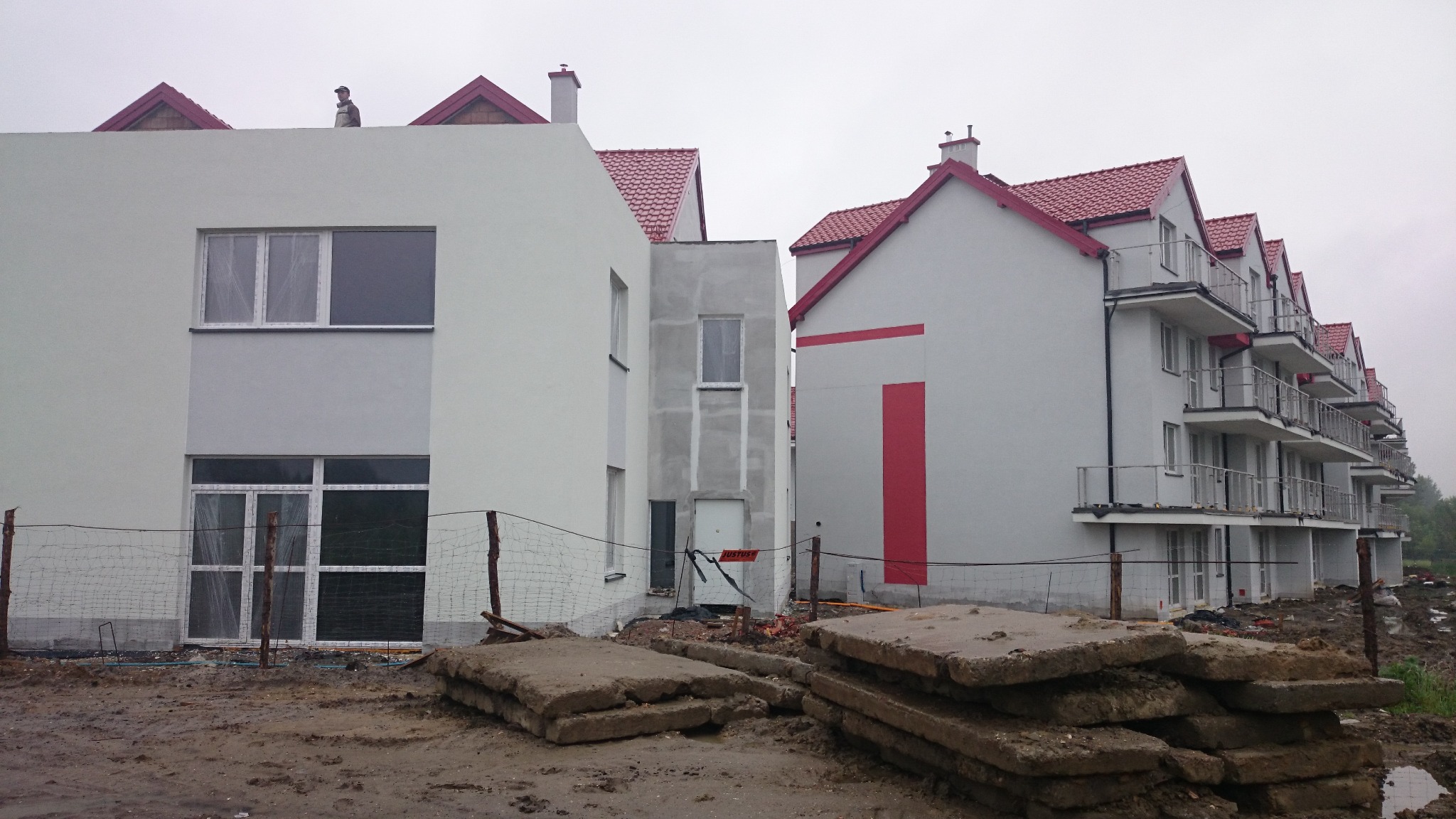 Wielorodzinny budynek w stanie deweloperskim z czerwonym dachem, widoczne betonowe płyty na błotnistym terenie i robotnik na dachu.