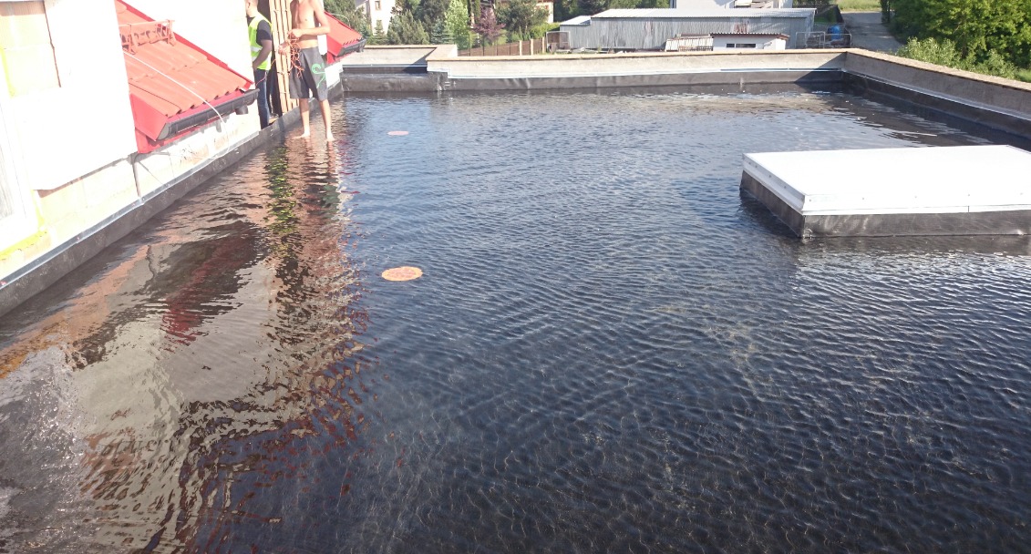 Płaski dach pokryty ciemną membraną, zalany wodą, z widocznymi pracownikami w tle i odbiciem czerwonego dachu budynku. Woda tworzy efekt lustra, odbijając elementy otoczenia.