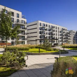 Nowoczesne osiedle mieszkaniowe z zadbanym dziedzińcem, zielenią i alejkami. Budynki w odcieniach szarości z balkonami. Słoneczny dzień, błękitne niebo.