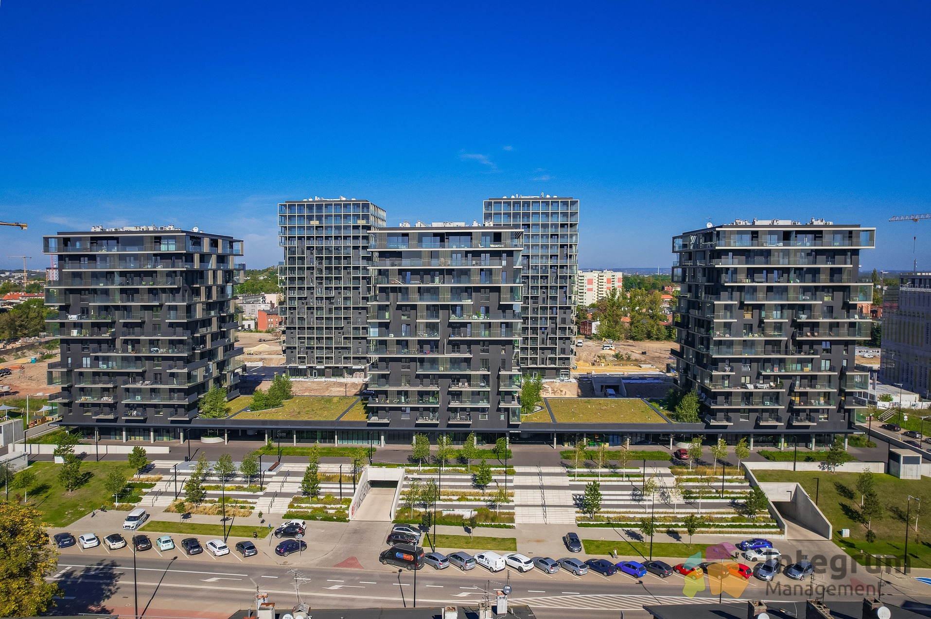 Nowoczesne apartamentowce z zielonymi dachami, parkingiem i uporządkowaną zielenią miejską w słoneczny dzień. Architektura z licznymi balkonami i prostą bryłą.